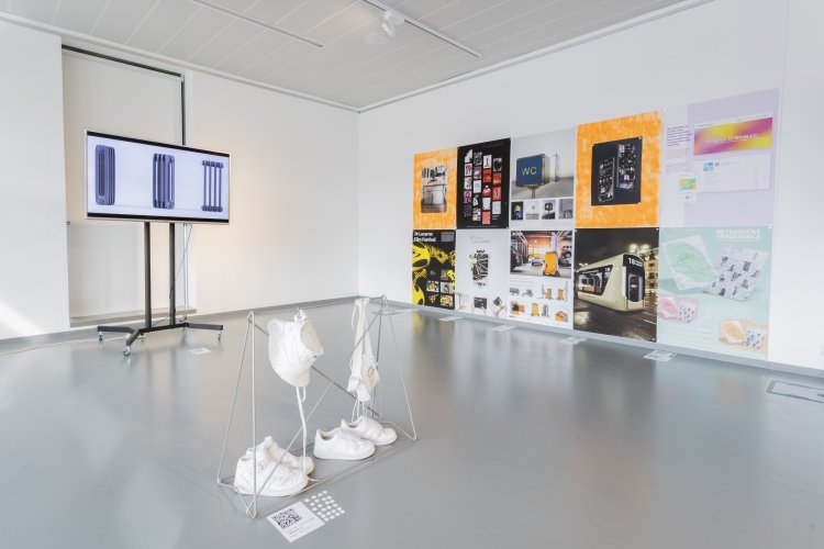 A view of the wall with 10 posters featuring projects from the Best in Design competition. On the left is a TV screen showing a radiator. In the foreground the installation is mounted on a metal stand. White sneakers are placed on the ground and a hat and a handbag, which are made just from upcycled sneakers, are hung on the metal stand.


