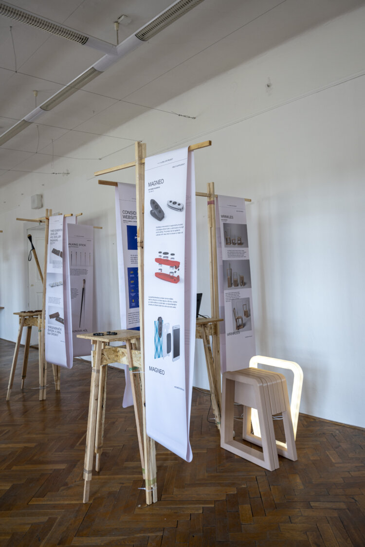 A view of the wooden installation with white banners presenting the exhibited projects. A wooden stool is placed next to the installation. The exhibition was titled long life and the projects on display are about upcycling and recycling.


