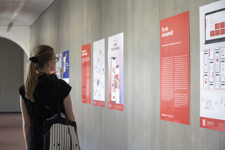 Mladší žena s vlasy v culíku si prohlíží výstavu věnují se tématice desinformací. Výstava je zpracovaná v podobě plakátů umístěných na stěnu. Plakáty mají červené pozadí a bílý text a jsou doplněny o grafické prvky.



